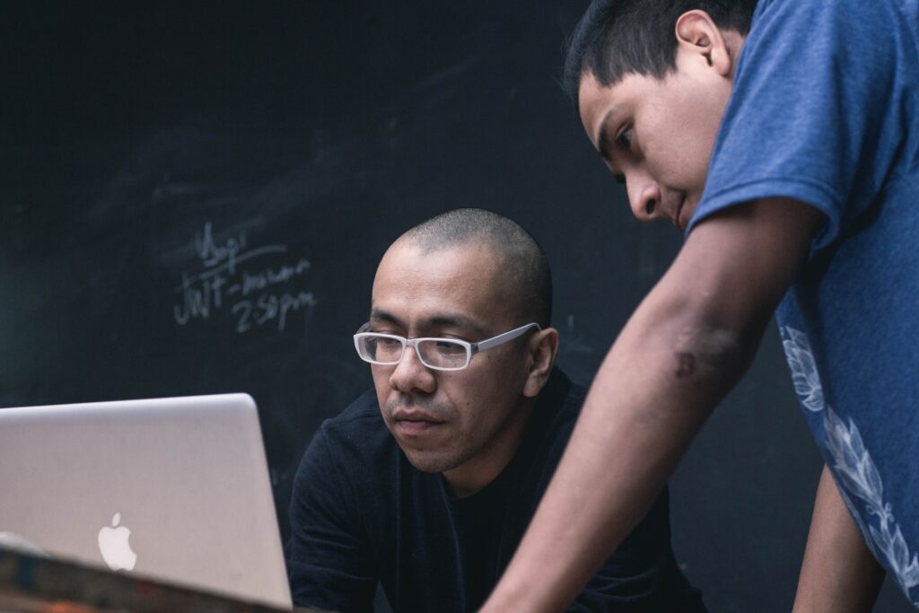 Two men looking at a laptop in front of a blackboard.