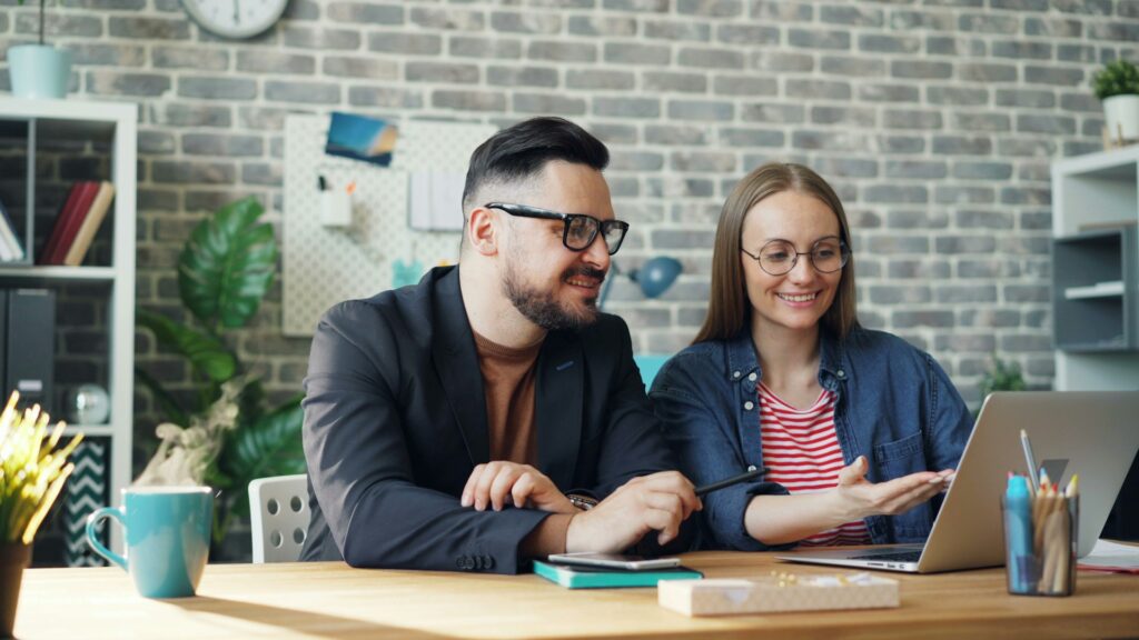 Male and female coworkers are discussing retrieval-augmented generation in finance while sitting at desk with laptop together and smiling.