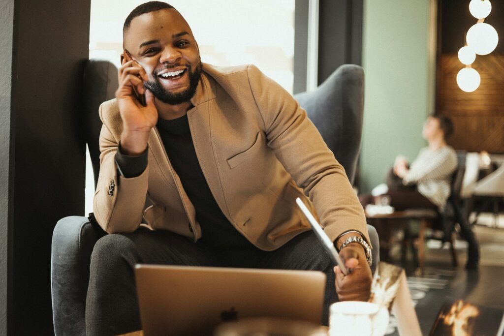 A man in a tan blazer sits on a leather chair, smiling and holding a laptop while talking on the phone about cloud-native.