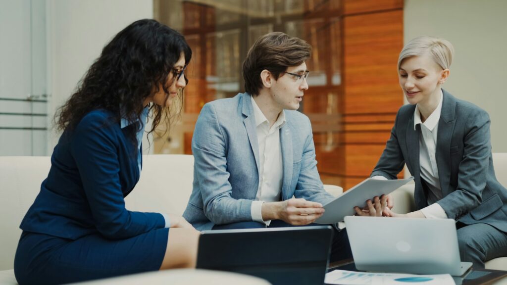 Three businesspeople in suits sit on a couch and discuss new technology adoption.