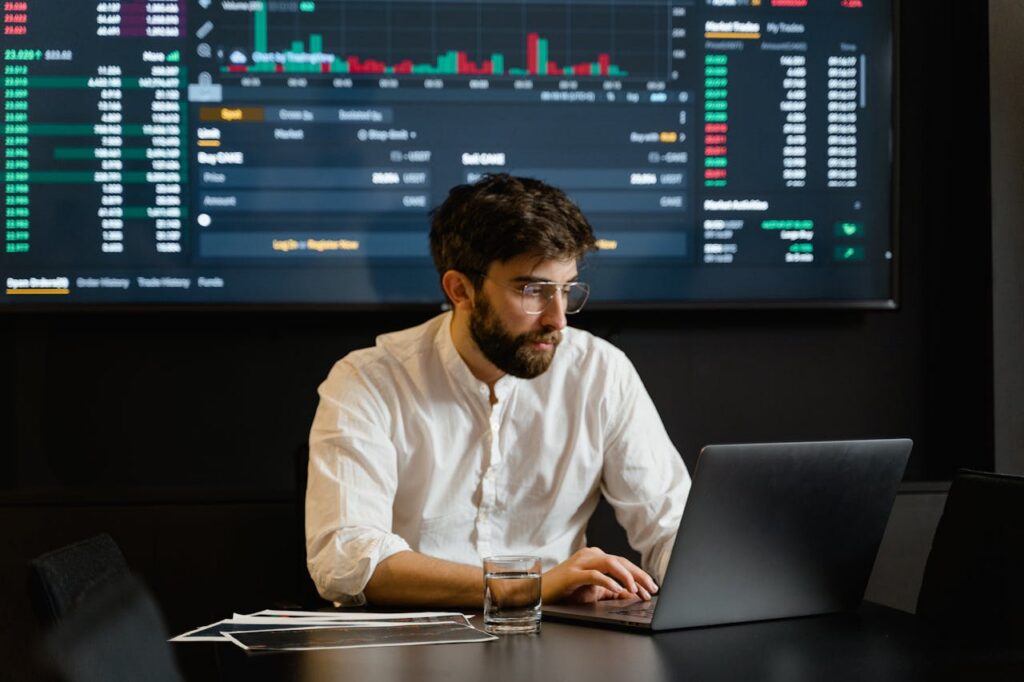 A bearded portfolio manager makes faster investment decisions in front of large screen depicting market price changes.