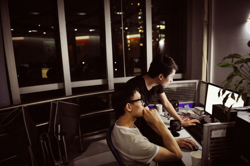Programmer colleagues modernize trading and risk systems at a desktop computer in an office at night.