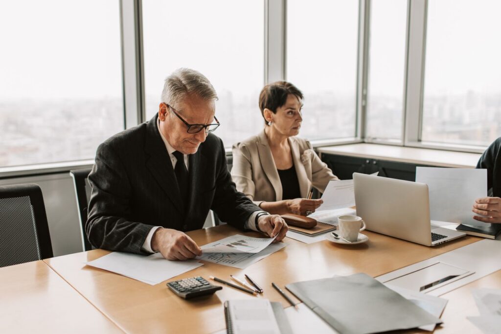 Stern, well-dressed executives sit at a conference table in a high-rise office, reviewing printed and digital reports on operational resilience.